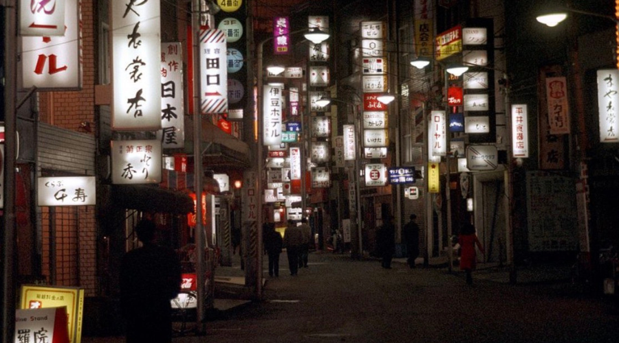 夜の繁華街に無数の看板が並ぶ路地を歩く人々の様子。素行調査や行動確認を連想させるシーン。